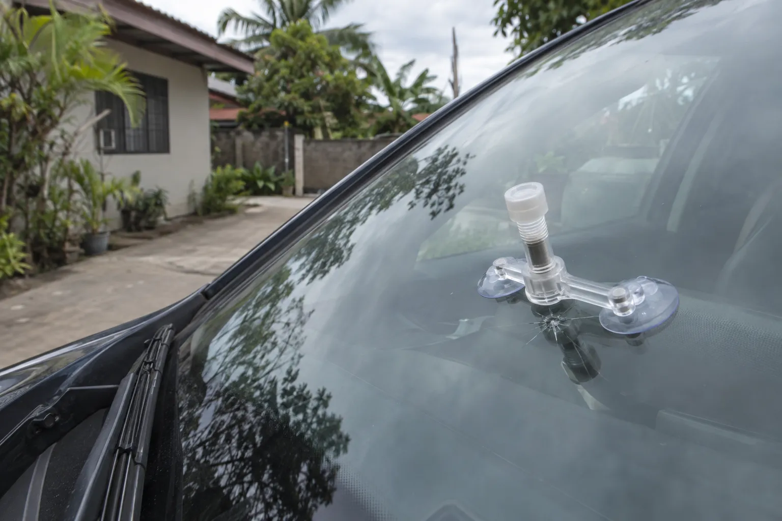 A resin bridge tool clamped over a small star chip on a windscreen, on a concrete driveway beside a Philippine house.