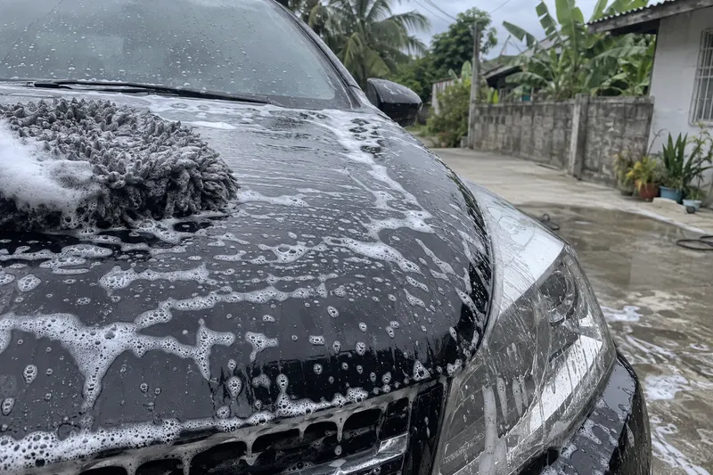 Wash foam worked across a dark bonnet with a mitt resting on it, on a driveway outside a house.