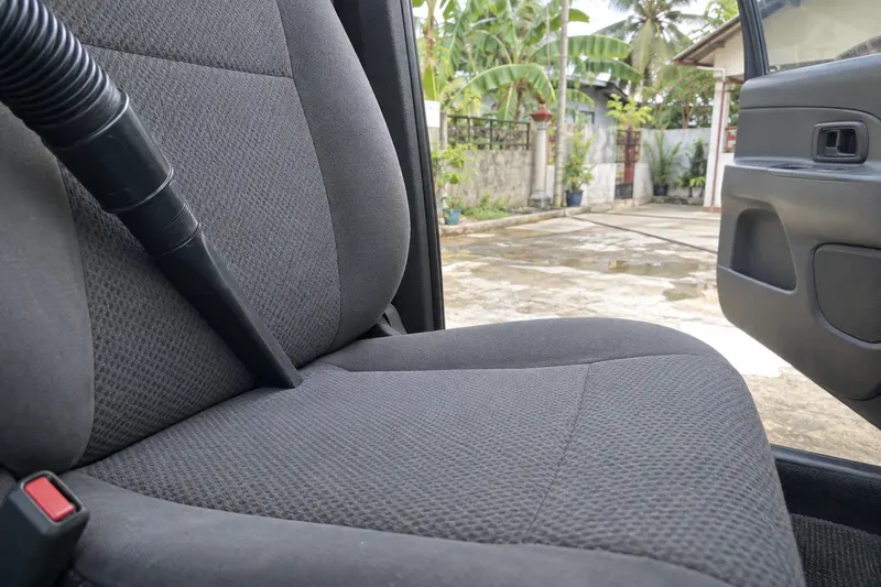 A vacuum nozzle pushed into the seam of a cloth car seat, the door open to a driveway.